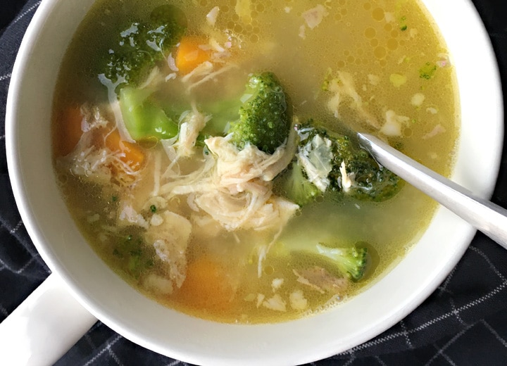 A spoon in a round white mug filled with broccoli, carrots, and shredded chicken in soup