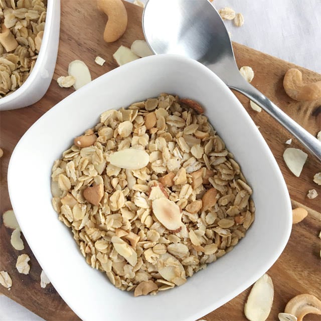 A spoon next to a square white bowl containing nutty granola