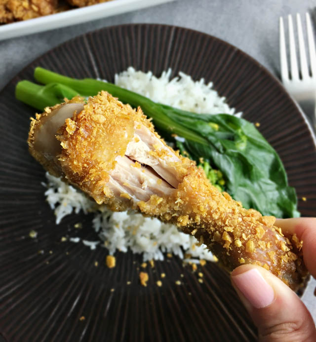 Fingers holding a cornflakes chicken drumstick with a bite taken out of it, a round plate with rice and green vegetables in the background