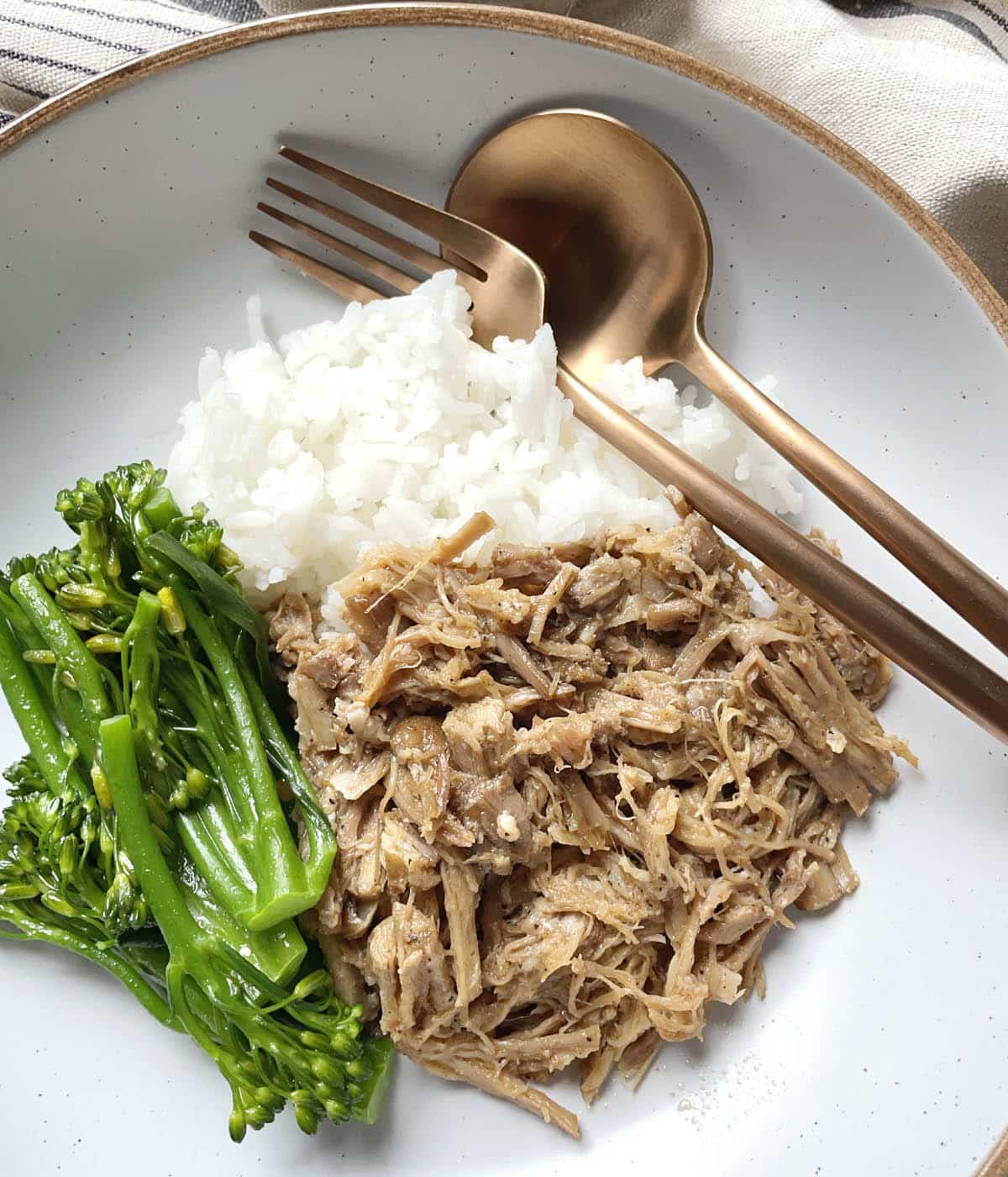 A round dish containing a fork and spoon, cooked white rice, green vegetables, and pulled pork.