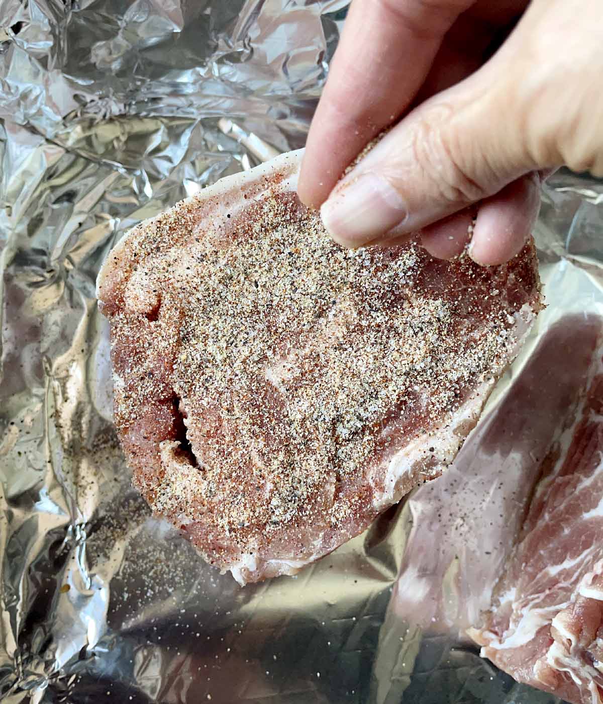 A hand sprinkling dry seasonings on a raw chunk of pork roast.