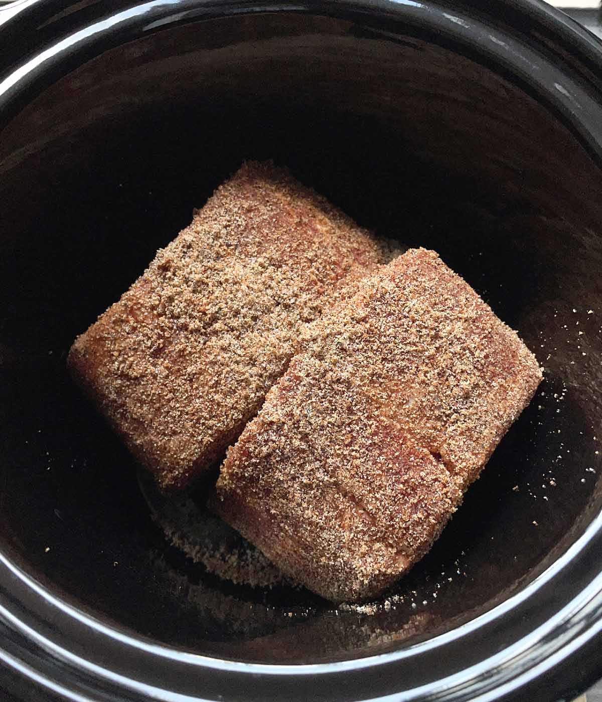 Two uncooked chunks of pork coated in dry seasonings in black pot.