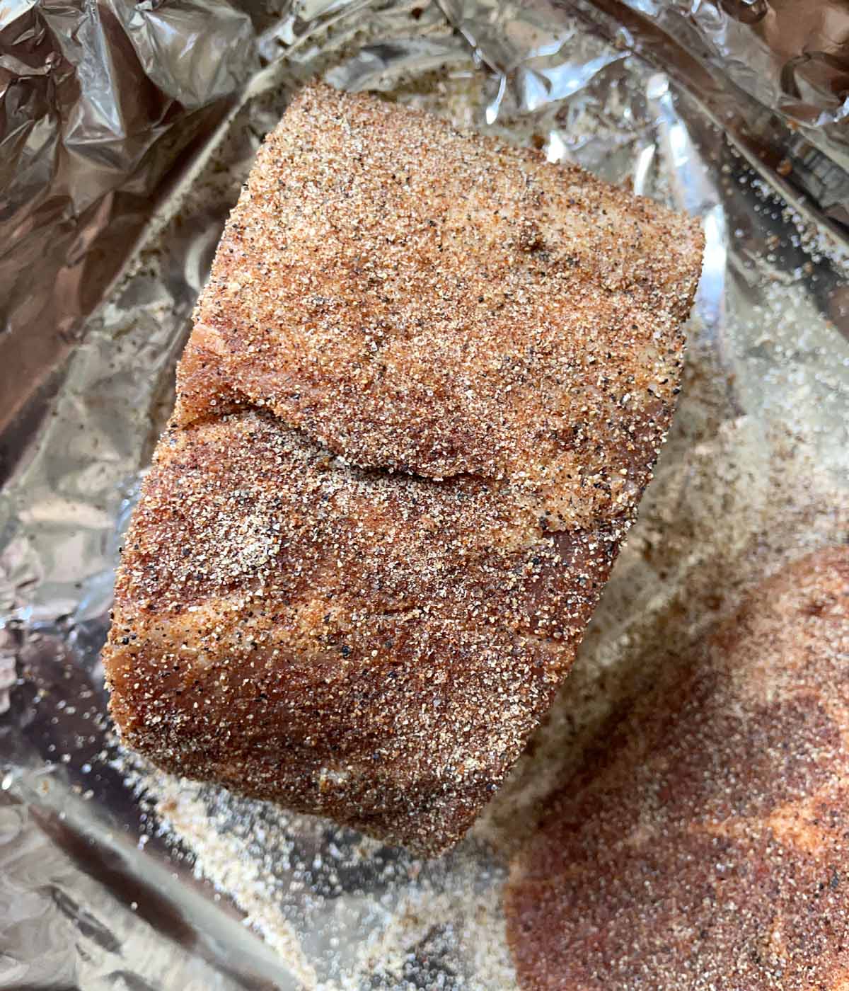 A raw chunk of pork coated in dry seasonings in a foil-lined pan.