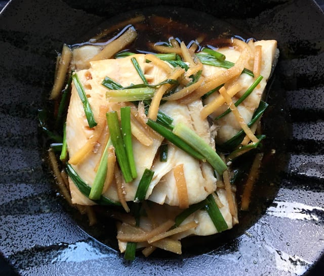 A black dish containing filets of fish and strips of green onion and ginger for poached black cod