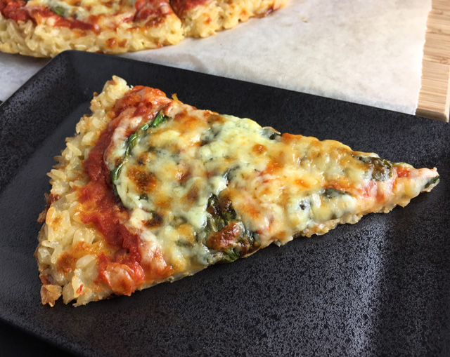 A black plate with a slice of Rice Crust Pizza made of rice, tomato sauce, spinach, and cheese