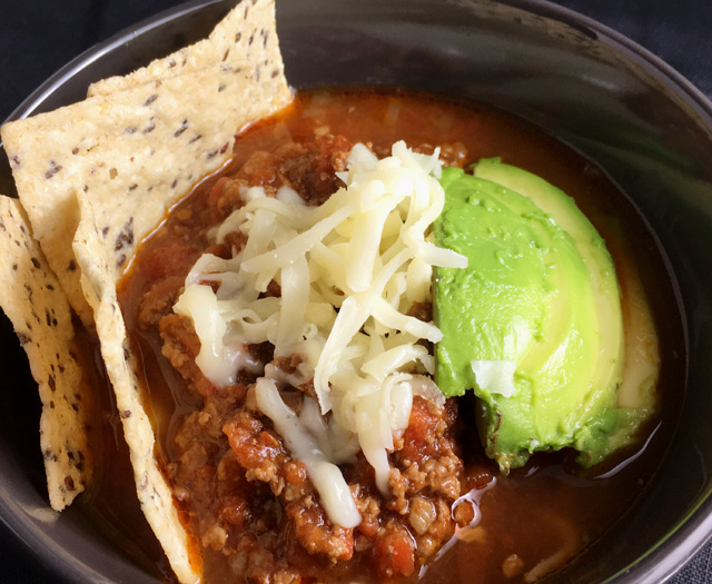A bowl of Chunky Chipotle Chili Con Carne with tortilla chips, avocado slices and grated cheese