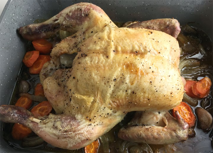 A whole roast chicken and carrots and onions in a grey roasting pan