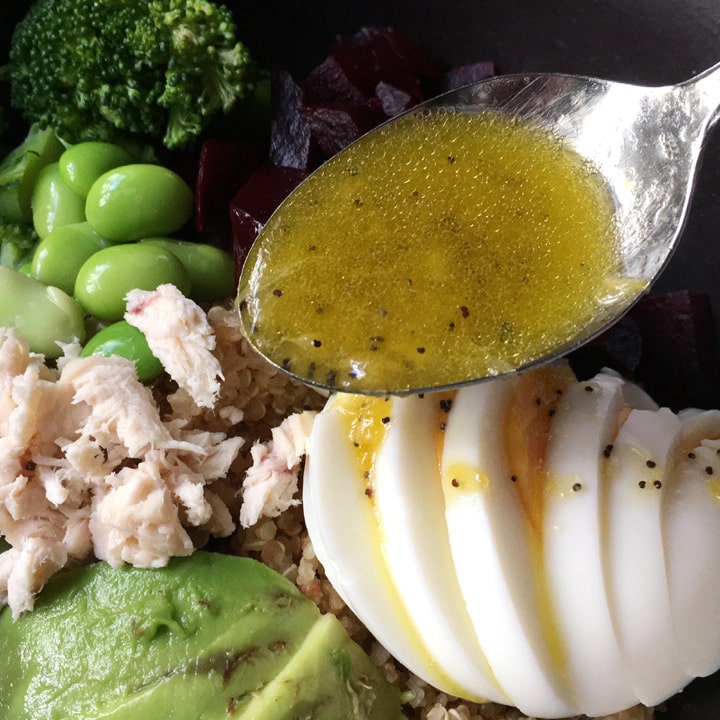 Close-up of a spoonful of orange dressing being drizzled over a bowl containing sliced egg, avocado, chicken, edamame beans