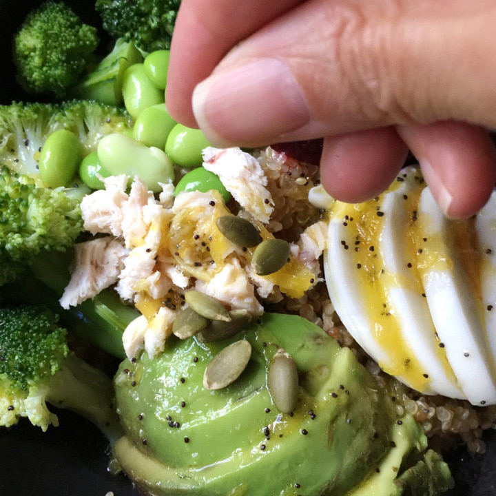 Close-up of a hand sprinkle pumpkin seeds over a bowl containing avocado, sliced egg, chopped chicken, edamame beans, and broccoli