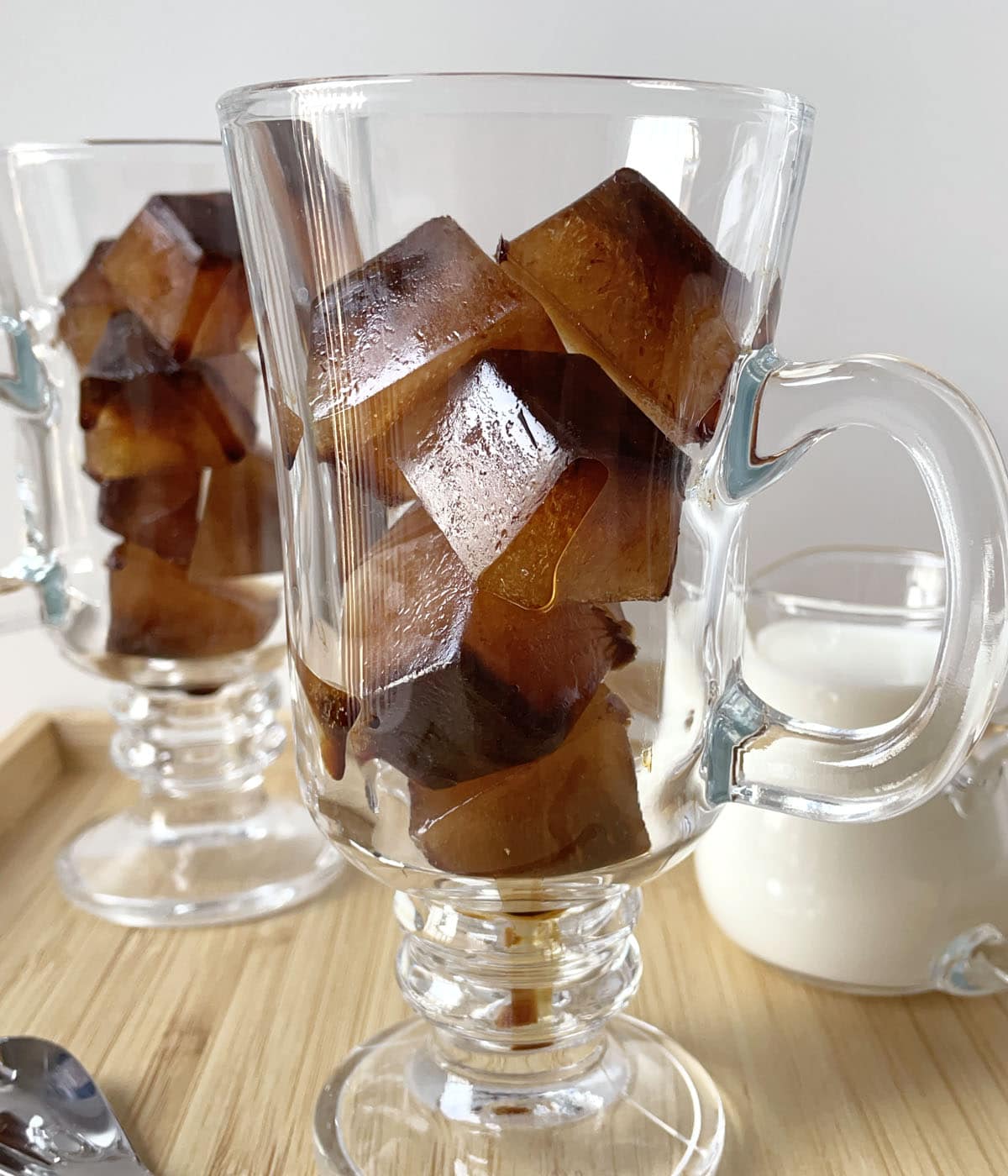 A small pitcher of milk next to two glass mugs containing coffee ice cubes.