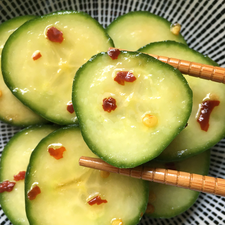 A pair of wood chopsticks picking up a thick cucumber slice with red chili flakes