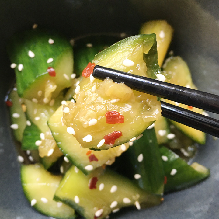 A pair of black chopsticks holding a crushed chunk of cucumber topped with white sesame seeds and red chili flakes