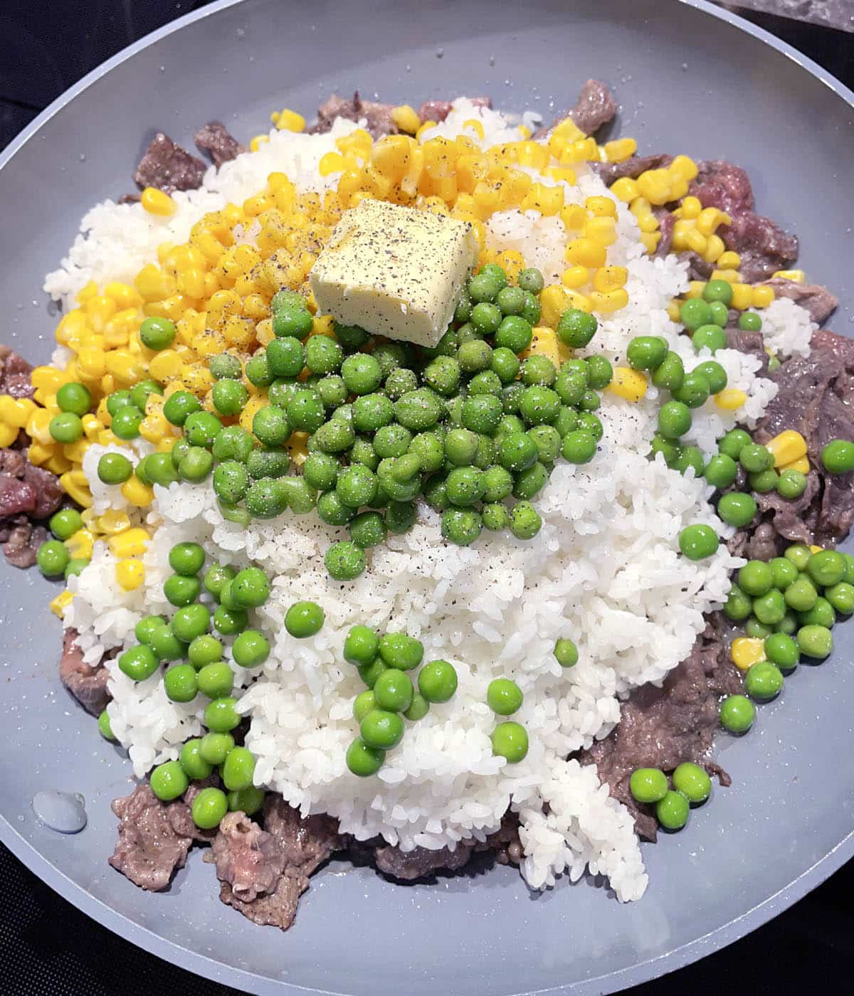 A round grey pan containing yellow corn, green peas, white rice, and beef.