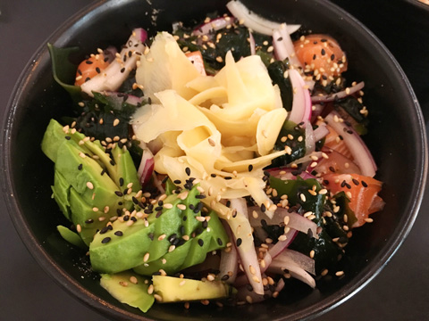 A black bowl containing rice, avocado, ginger slices, onions, raw salmon, seaweed, and sesame seeds at Poke HK