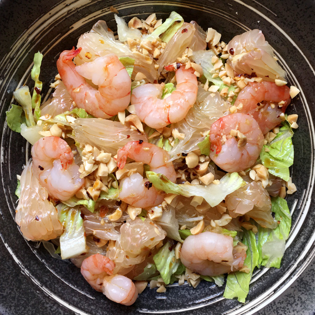 A dark metal bowl containing chopped lettuce, pomelo wedges, pink shrimp, and chopped peanuts