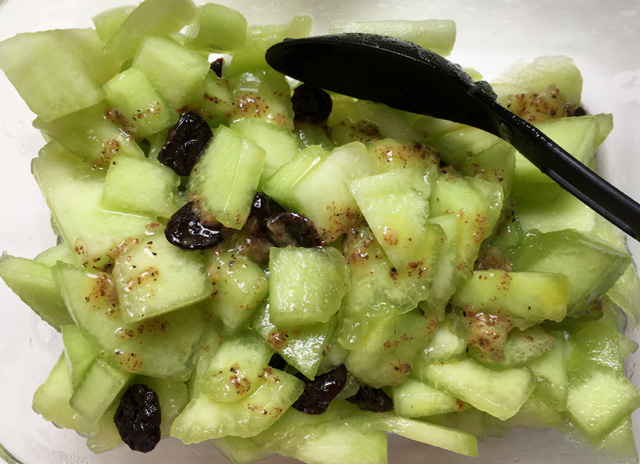 A bowl containing cut-up honeydew, dried cranberries, and dressing for Pistachio Cranberry Melon Salad