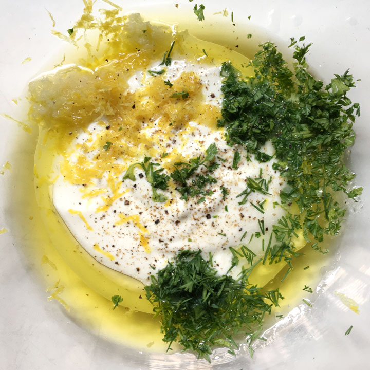 A glass bowl containing white yogurt, chopped green herbs, yellow lemon zest, yellow oil, ground black pepper