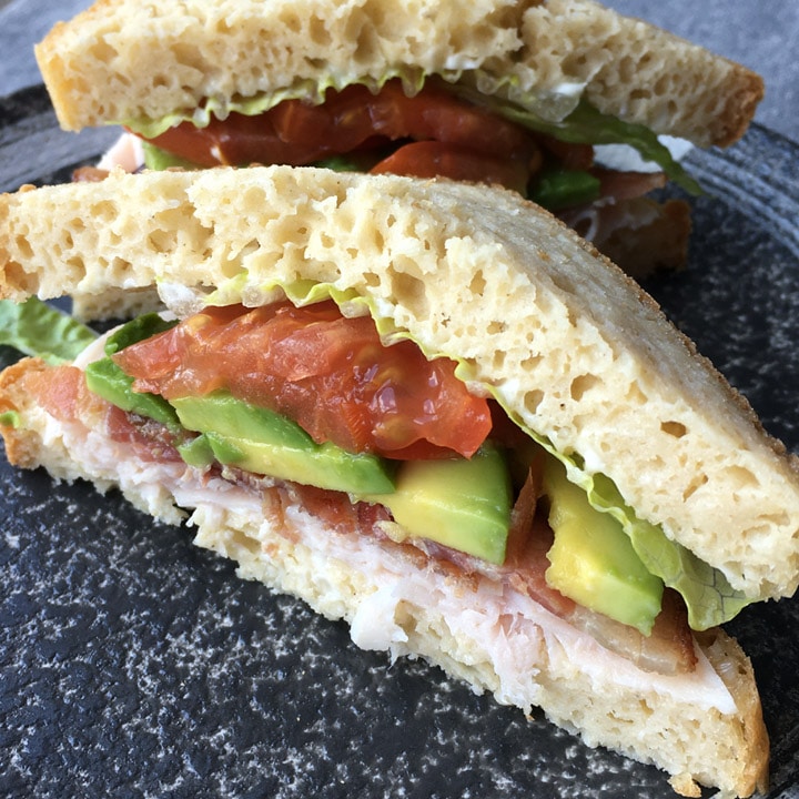 A round plate containing a sandwich filled with red tomatoes, green avocado, white meat