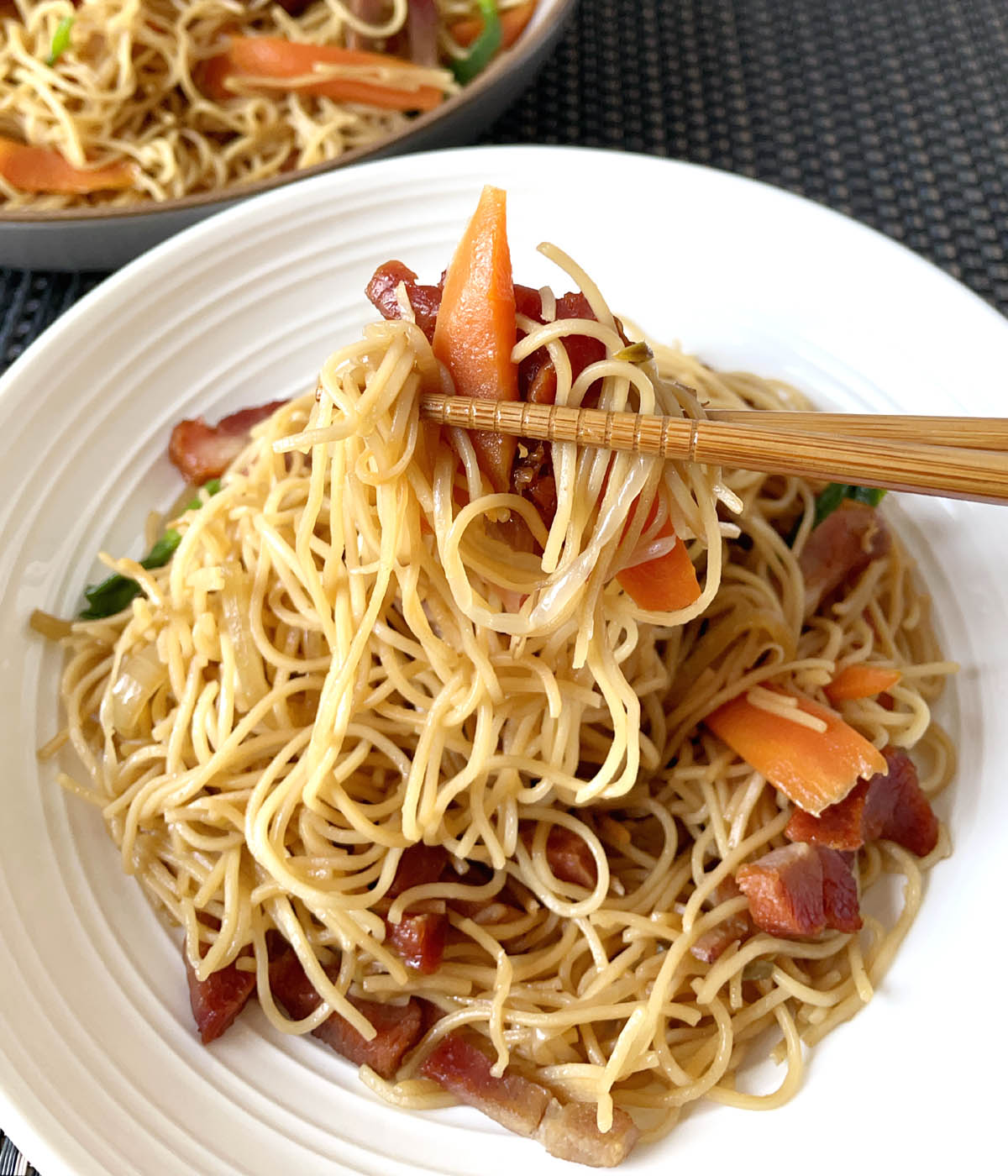 A pair of wooden chopsticks holding up  noodles, carrots and meat over a white round dish.