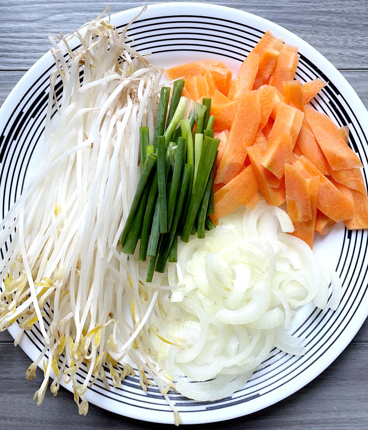 A round white plate containing bean sprouts, green onions, sliced white onions, and carrots.