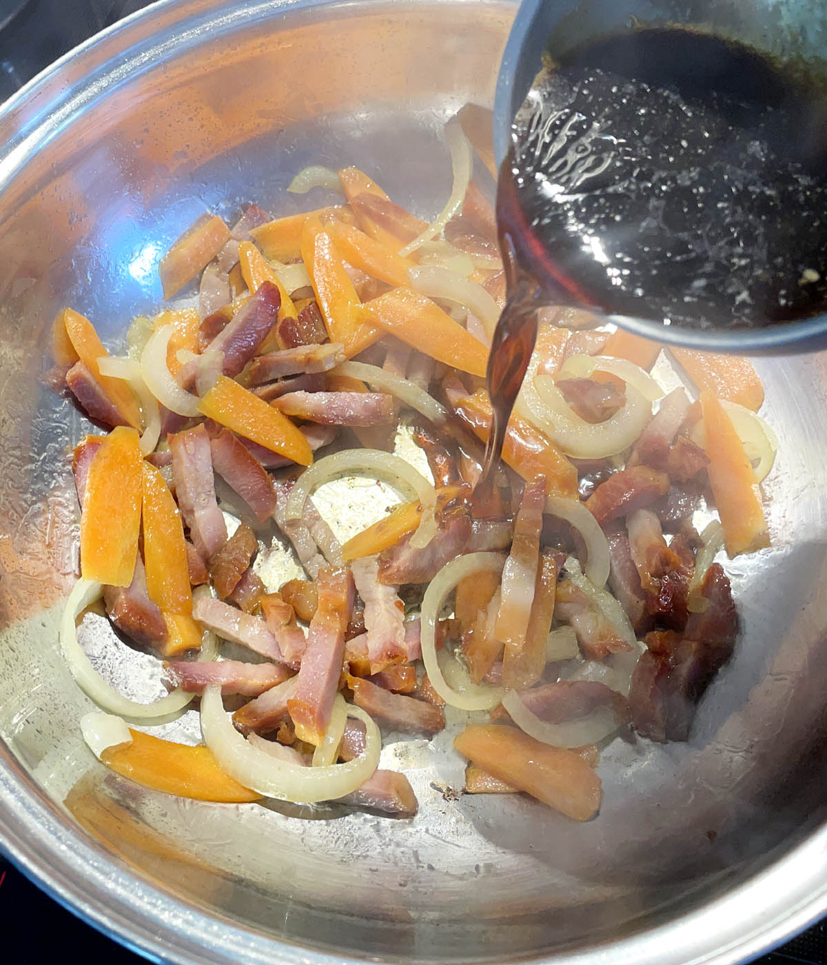 Brown liquid being poured into a wok containing sliced onions and carrots and chunks of meat.
