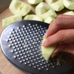 A hand holding a peeled apple chunk on a metal grater, chunks of apple in the background for flourless chocolate chocolate chip muffins