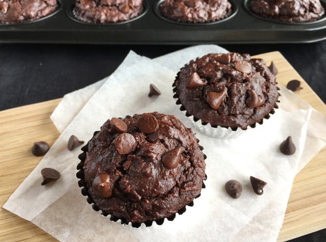 Two flourless chocolate chocolate chip muffins and chocolate chips on white parchment paper on a wooden cutting board, with a tray of muffins in the background