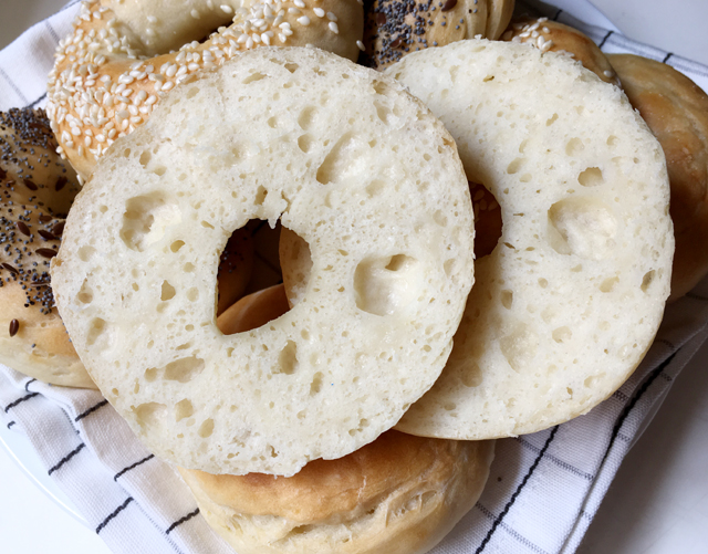 A sliced homemade bagel resting on a pile of baked bagels in a white napkin-lined dish