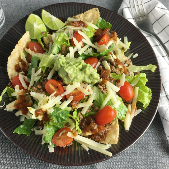 A dark brown plate containing tortilla chips, green lettuce, red tomatoes, ground beef, and cheddar cheese for taco salad