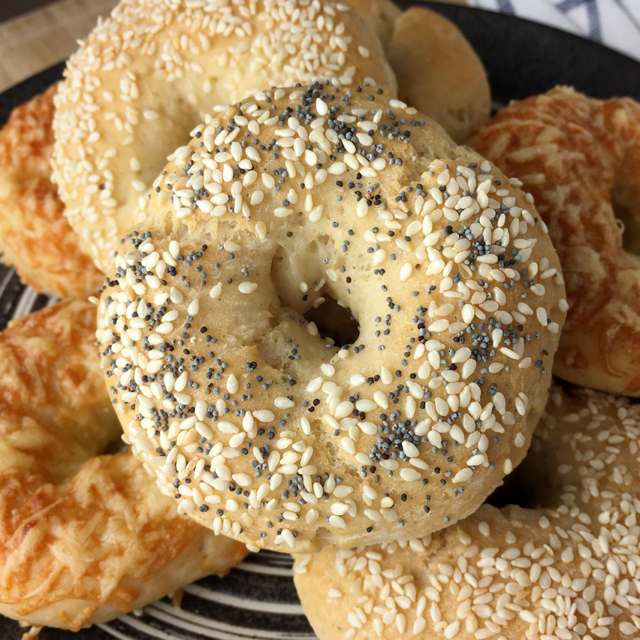 A bagel topped with sesame seeds and poppy seeds, on top of a pile of gluten-free bagelss on a plate