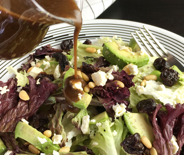 Balsamic vinaigrette being poured over a salad of avocado, feta, pine nuts, and lettuce greens