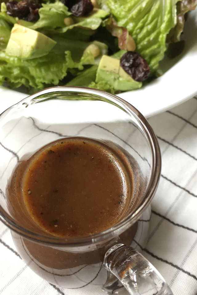 A closeup of balsamic vinaigrette in a glass pitcher next to a salad in a white plate