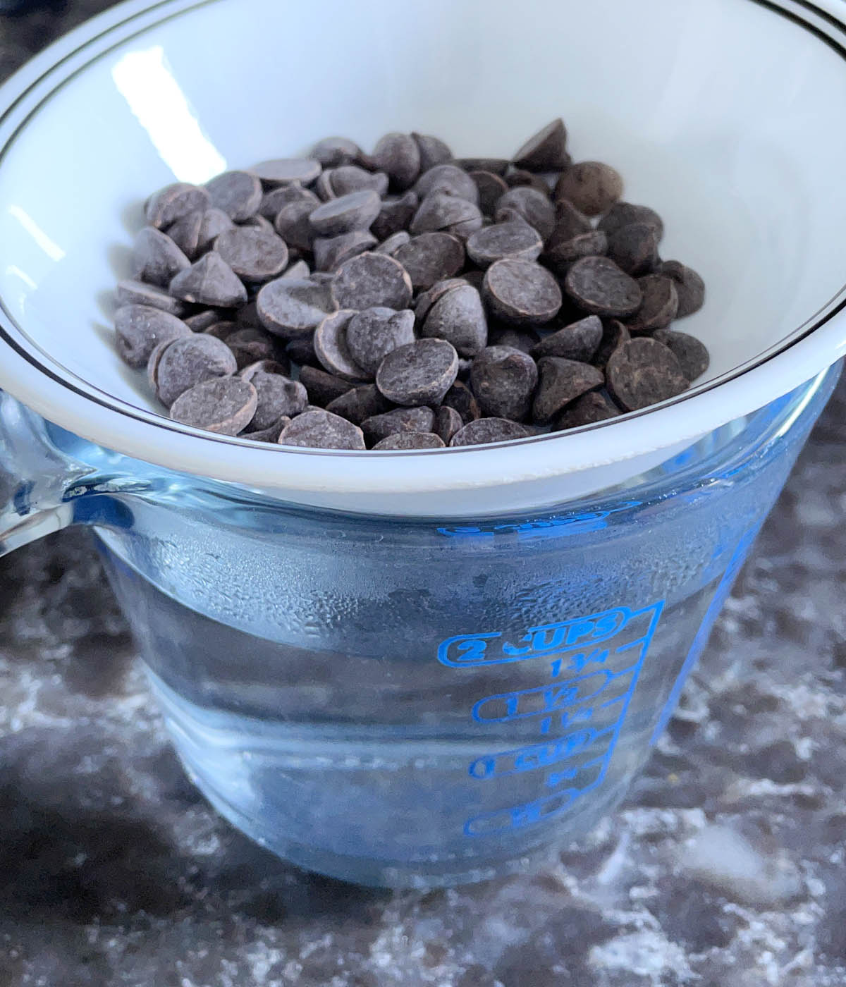 A white bowl containing chocolate chips over a glass measuring cup filled with hot water.