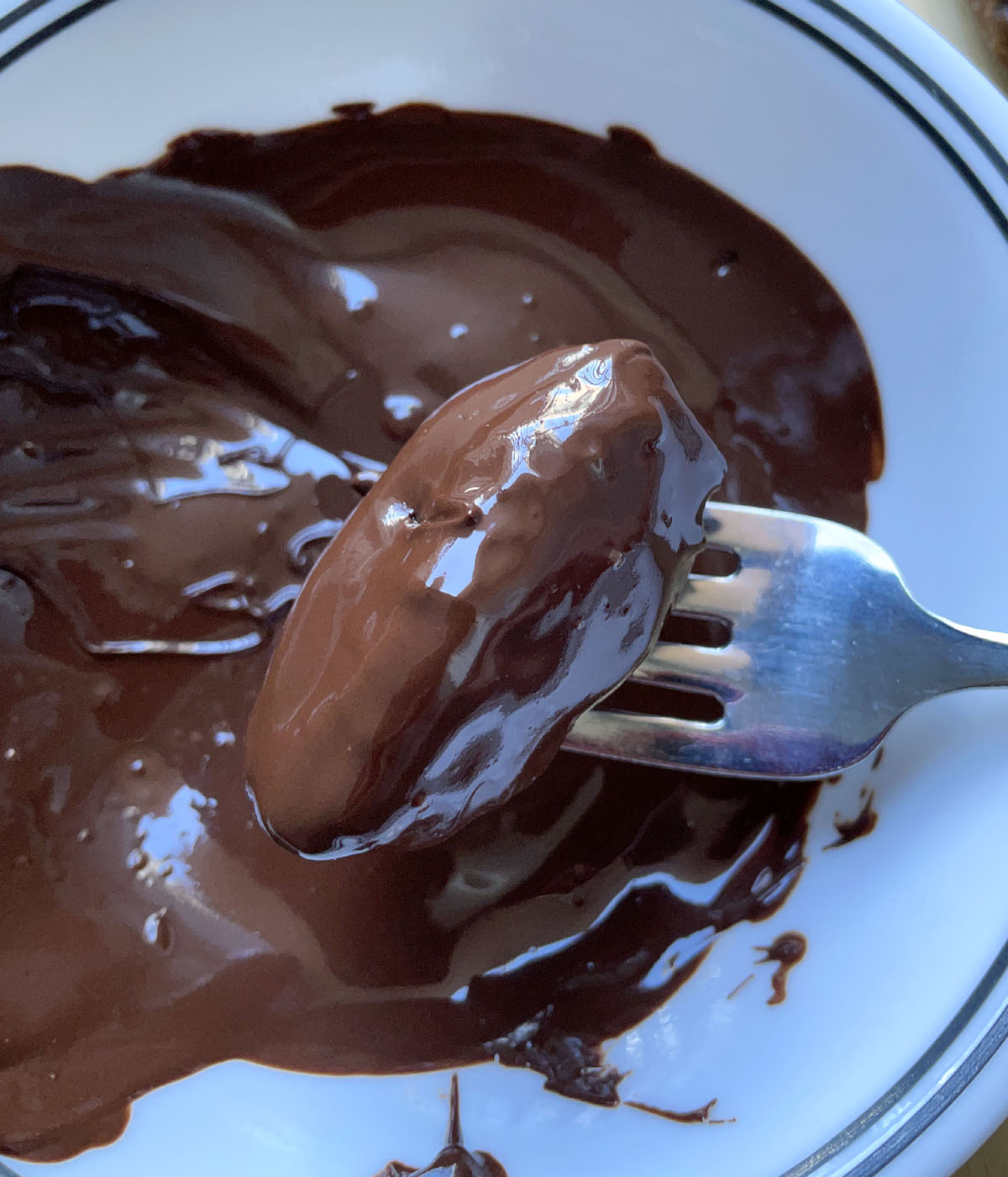 A fork holding a chocolate coated date over a white bowl of melted chocolate.