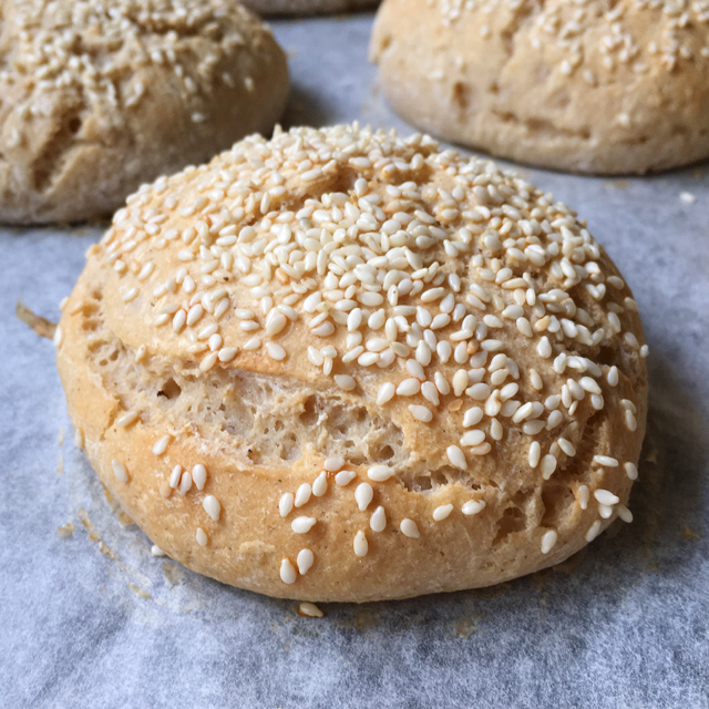 Close-up of a gluten-free bun, topped with sesame seeds and sitting on white parchment paper