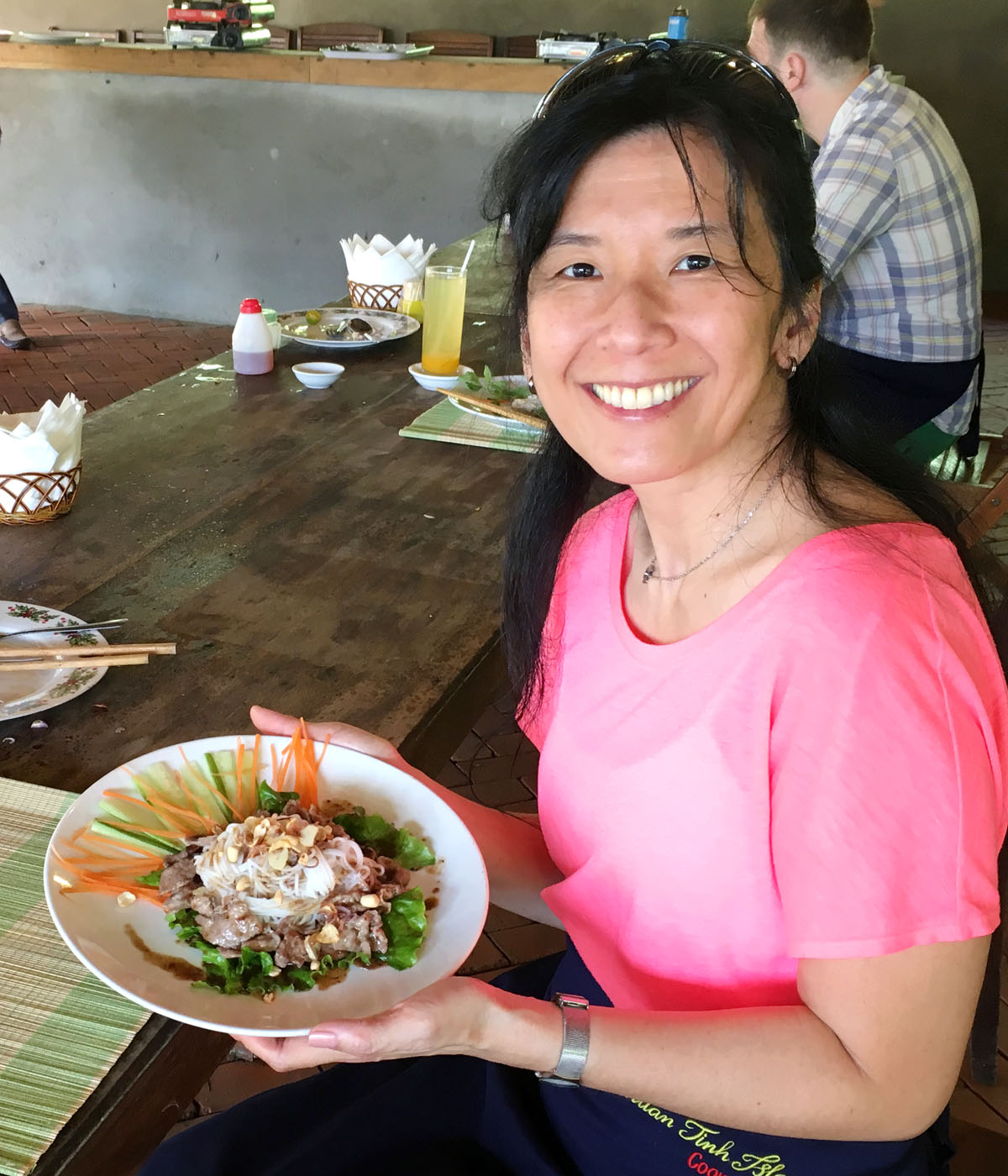 Asian woman wearing a pink shirt, holding a white dish containing vegetable and noodles.