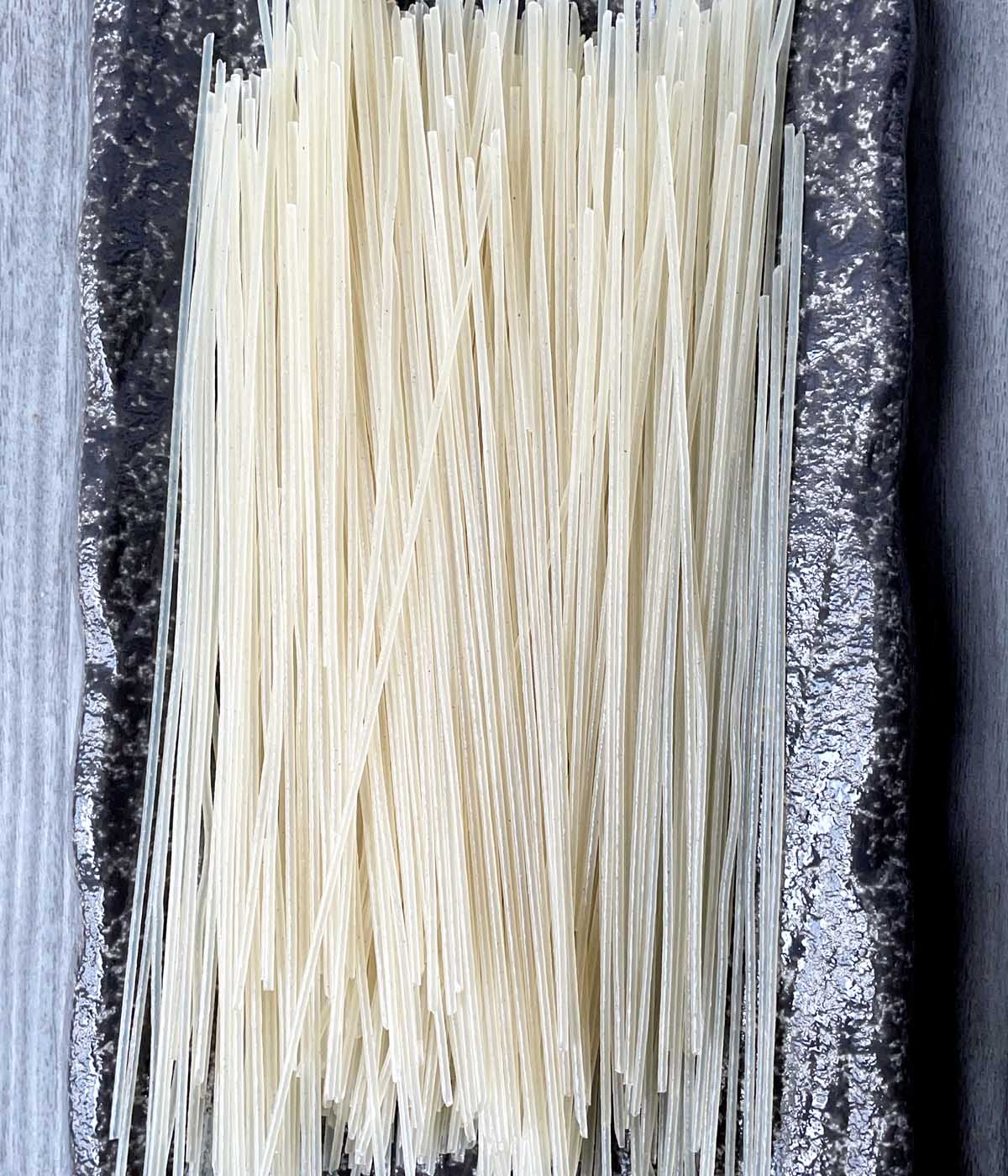 A dark grey rectangular plate containing white uncooked stick noodles.