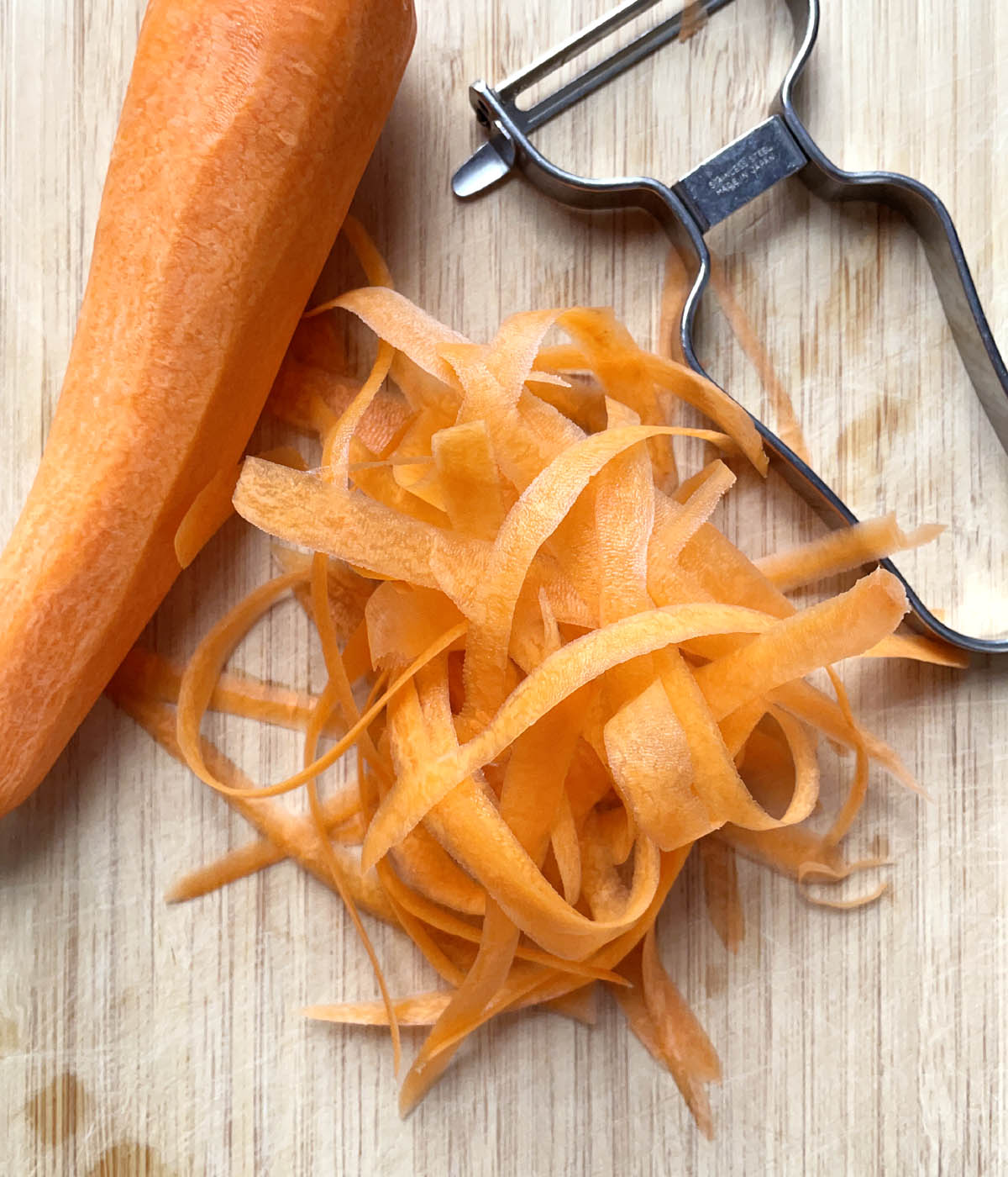 A carrot, a vegetable peeler, and carrot peelings on a wooden cutting board.
