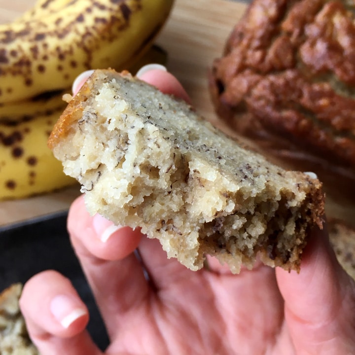 Close-up of a hand holding a piece of banana bread, yellow bananas in the background