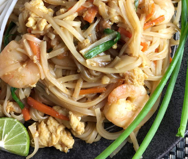 Close-up of a plate of shrimp pad thai with green onion stalks, cut lime, and white bean sprouts