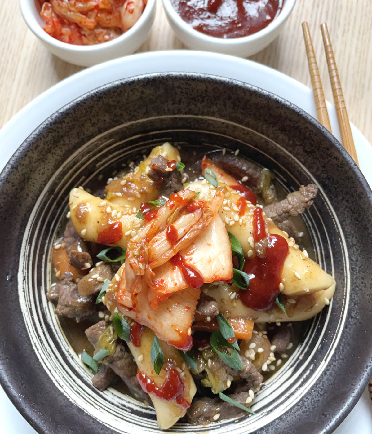 A dark round dish containing stir-fried rice cakes, cooked beef and vegetables, and topped with kimchi and red chili sauce.