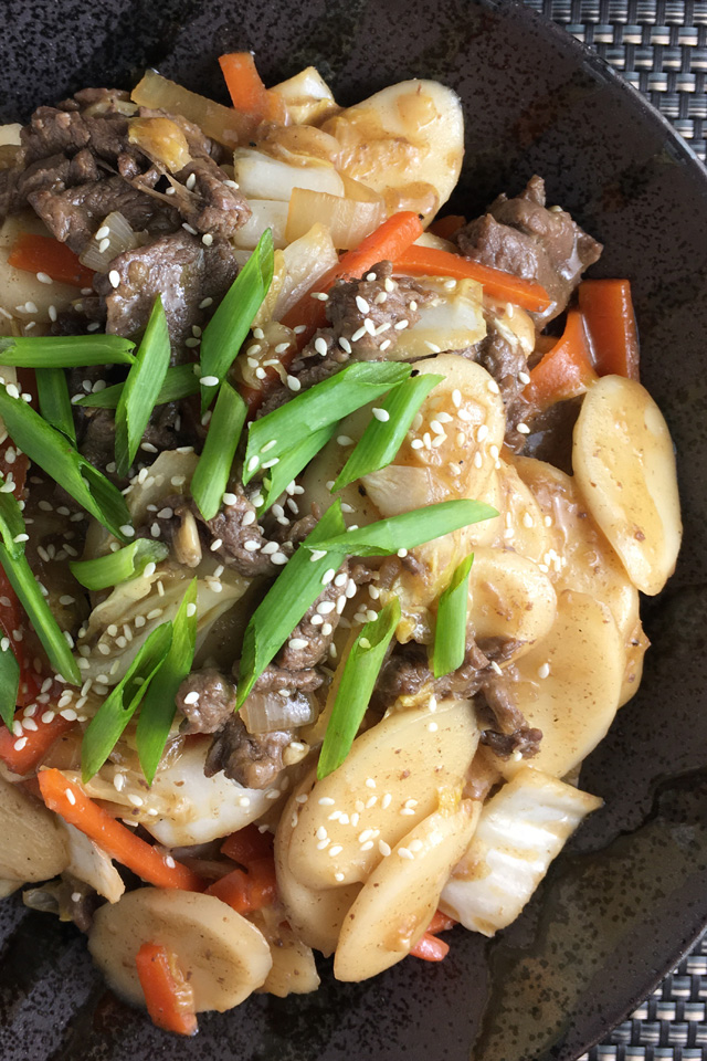 Close-up of a black dish containing stir-fried rice cakes with beef, carrots, cabbage, green onions, and sesame seeds