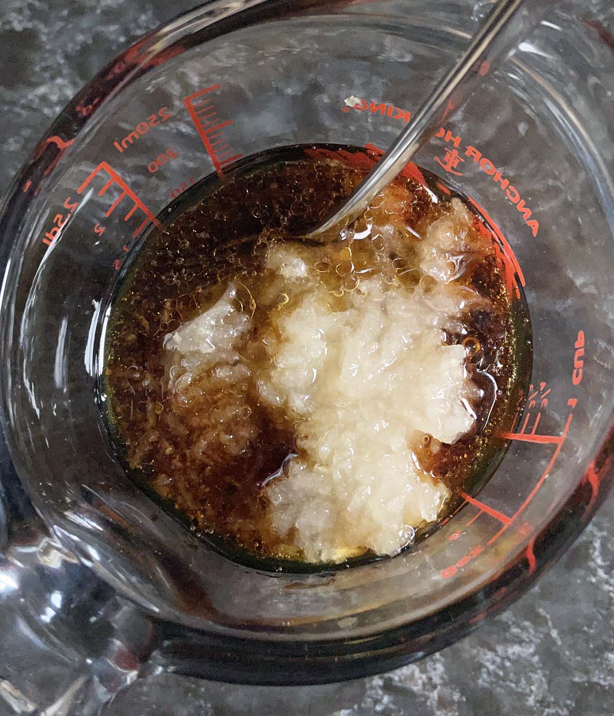 A glass measuring cup containing a metal spoon, brown liquid and grated pear.