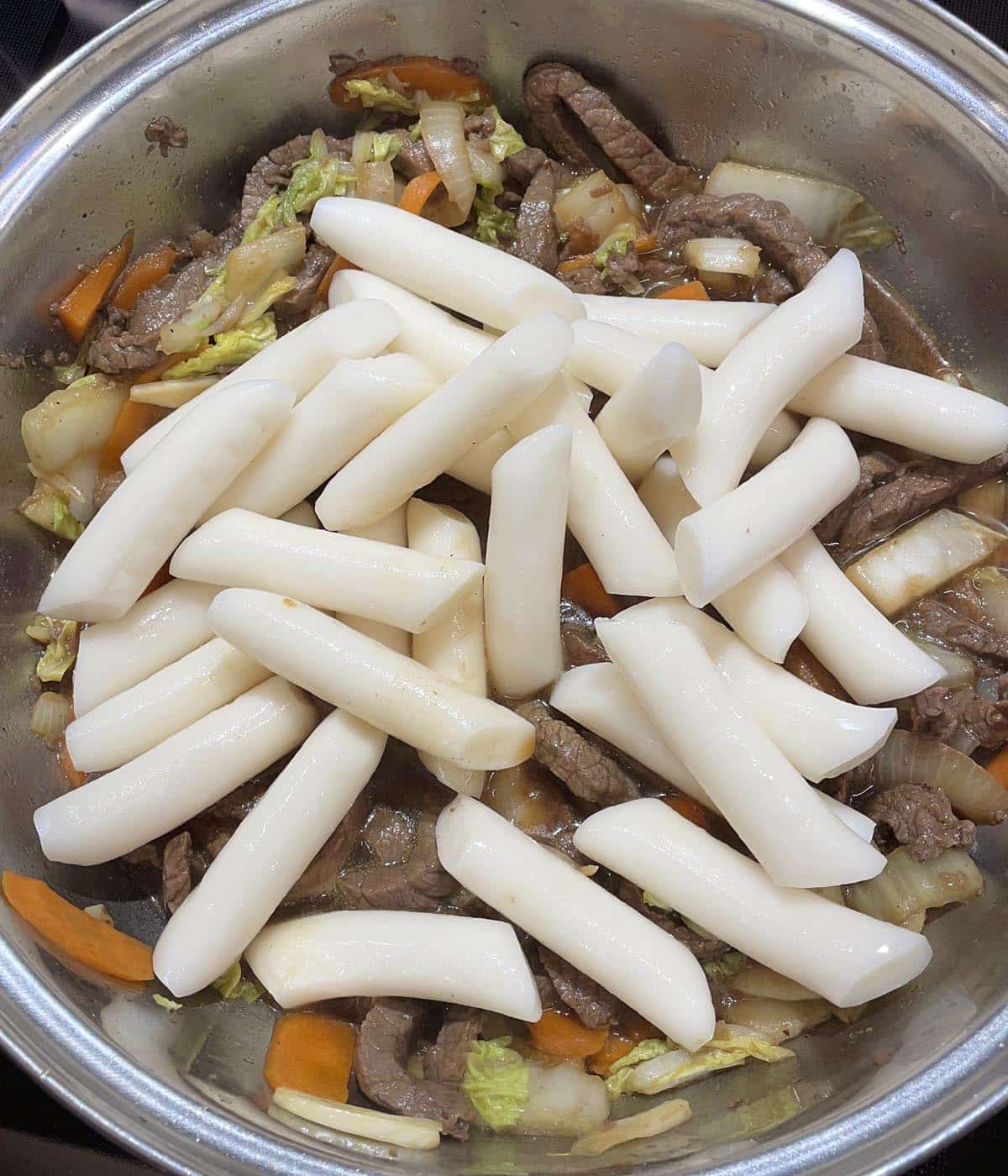 White cylinder shaped rice cakes in a metal pan containing cooked meat and vegetables.