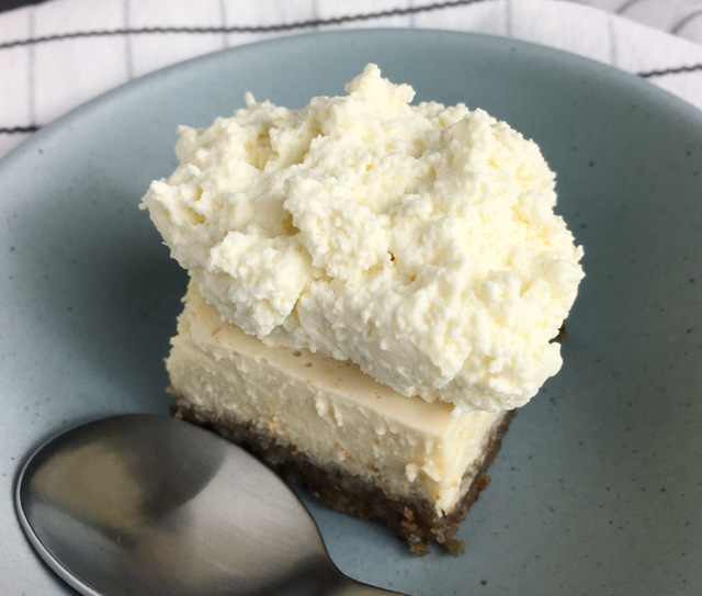A cheesecake square with whipped cream on top in a light blue dish with a spoon