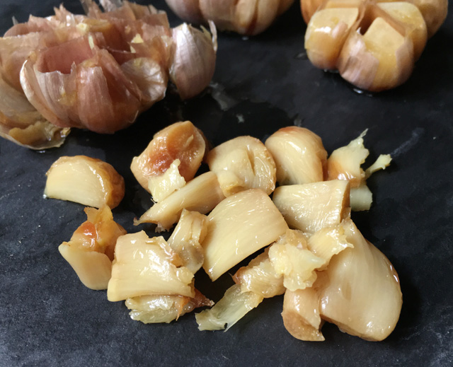 Roasted garlic on a grey stone surface with an empty garlic bulb in the background