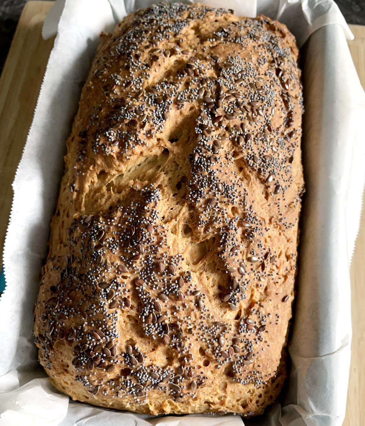 A brown loaf of bread topped with flax seeds and poppy seeds, in a paper lined baking pan.