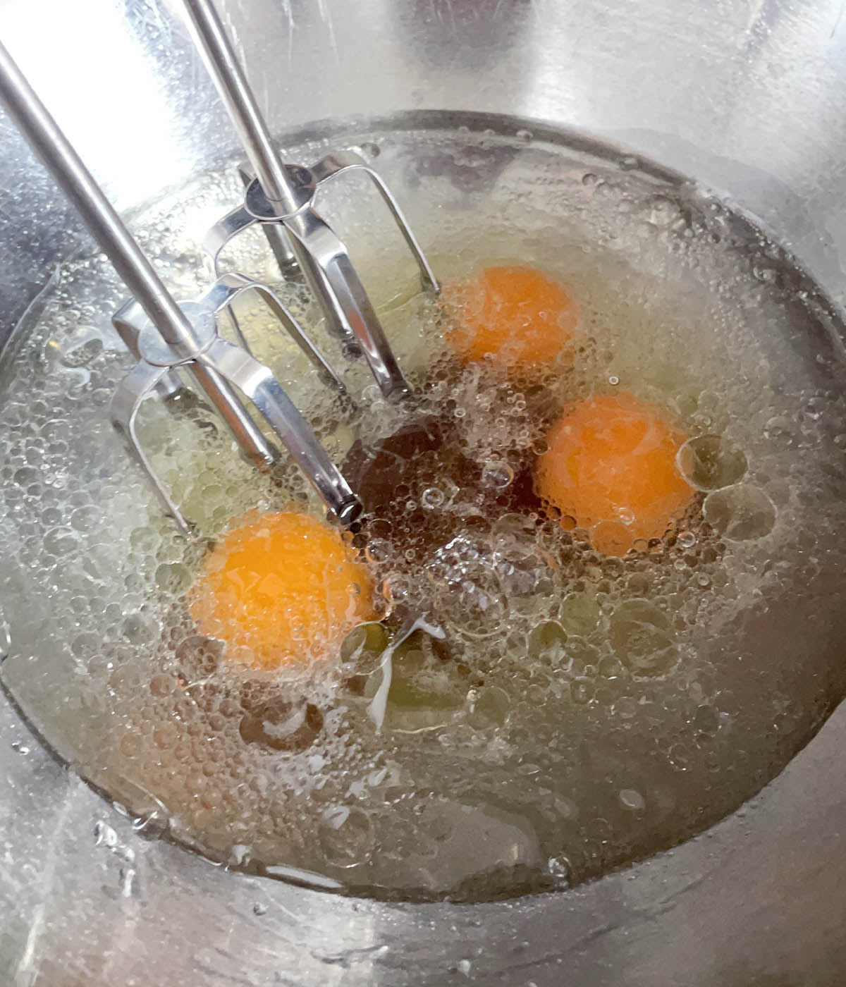 Two mixer beaters in a metal round bowl containing 3 egg, oil, and clear liquid.