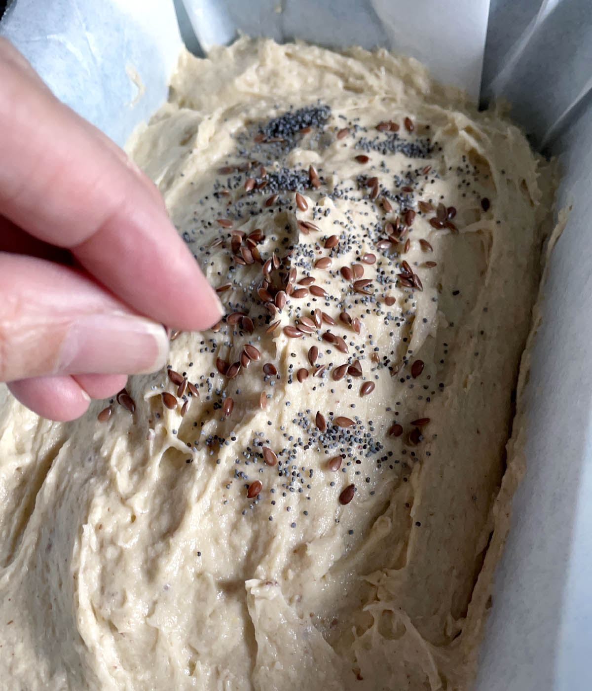 A hand sprinkling brown flax seeds and grey poppy seeds over dough in a bread pan.
