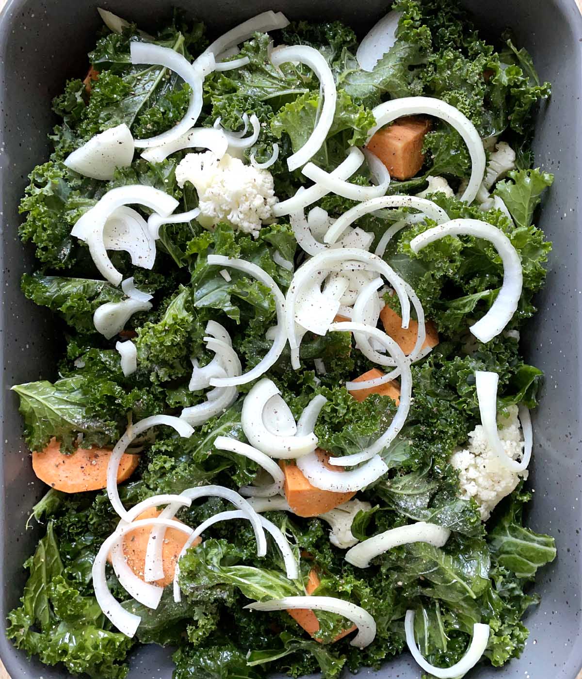 A rectangular roasting pan containing uncooked green kale, white cauliflower, orange sweet potatoes, and thinly sliced onions.